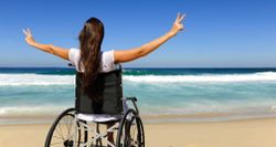 Stock photo of woman in wheelchair one beach with arms raised to heaven and hands making peace signs.