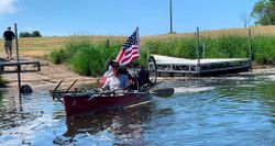 Man on canoe departing river launch point, with large American flag positioned at back of canoe.
