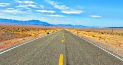 Two-lane road leading through desert-like area and toward mountains in the distance.
