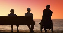 Two people sitting on a bench and one wheelchair user watching the sunset.