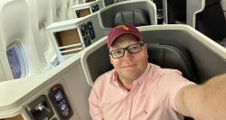Selfie of John wearing a pink shirt and garnet baseball cap seated in business class on a Boeing 777 aircraft.
