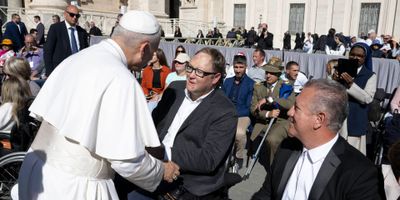 John seated in his power wheelchair shaking hands with Pope Leo.