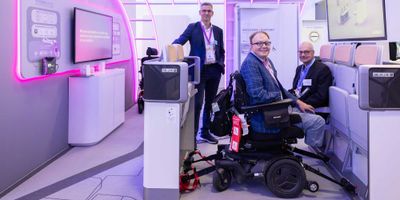 John seated in a wheelchair securement space on the Airbus stand, posing with two Airbus employees.