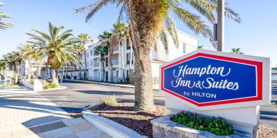 Hampton Inn and Suites sign in front of hotel with palm trees and sidewalk.