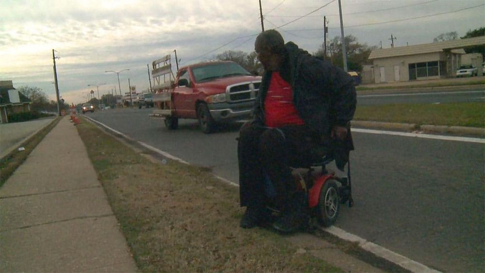 A man is forced to ride his wheelchair in the road because the sidewalk is not accessible.