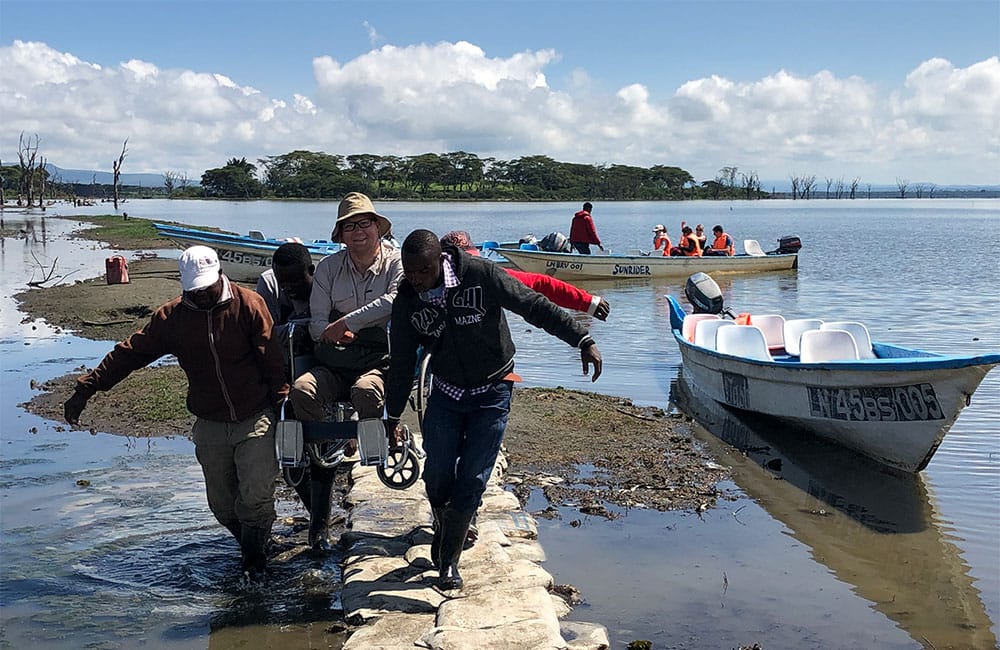 4 men carrying wheelchair across water.