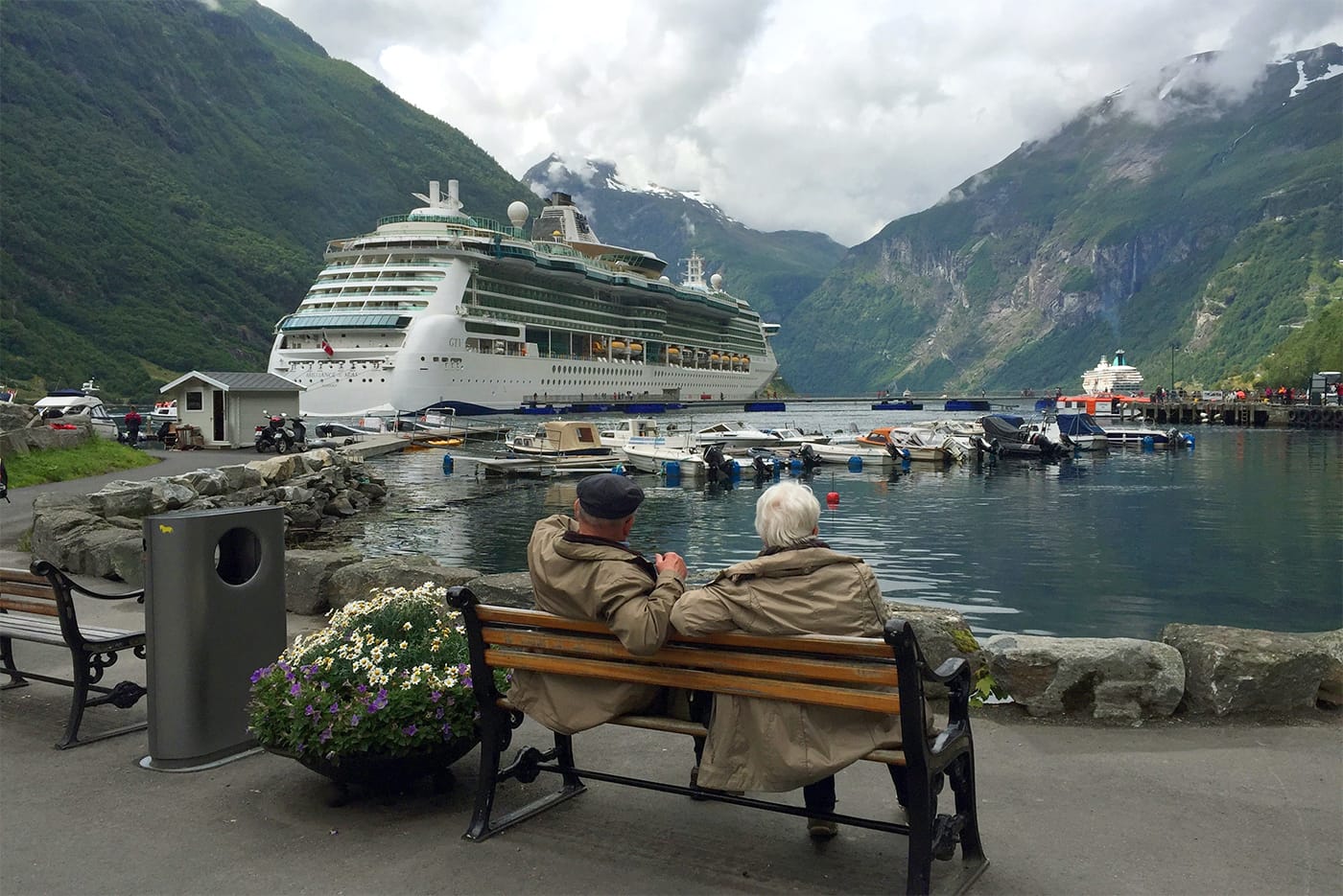 Elderly couple sitting on a bench looking at a cruise ship docked at port on an overcast day.