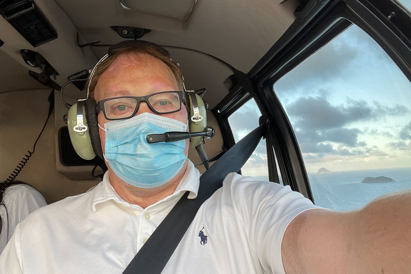 John seated in the helicopter wearing a headset, seatbelt and face mask.