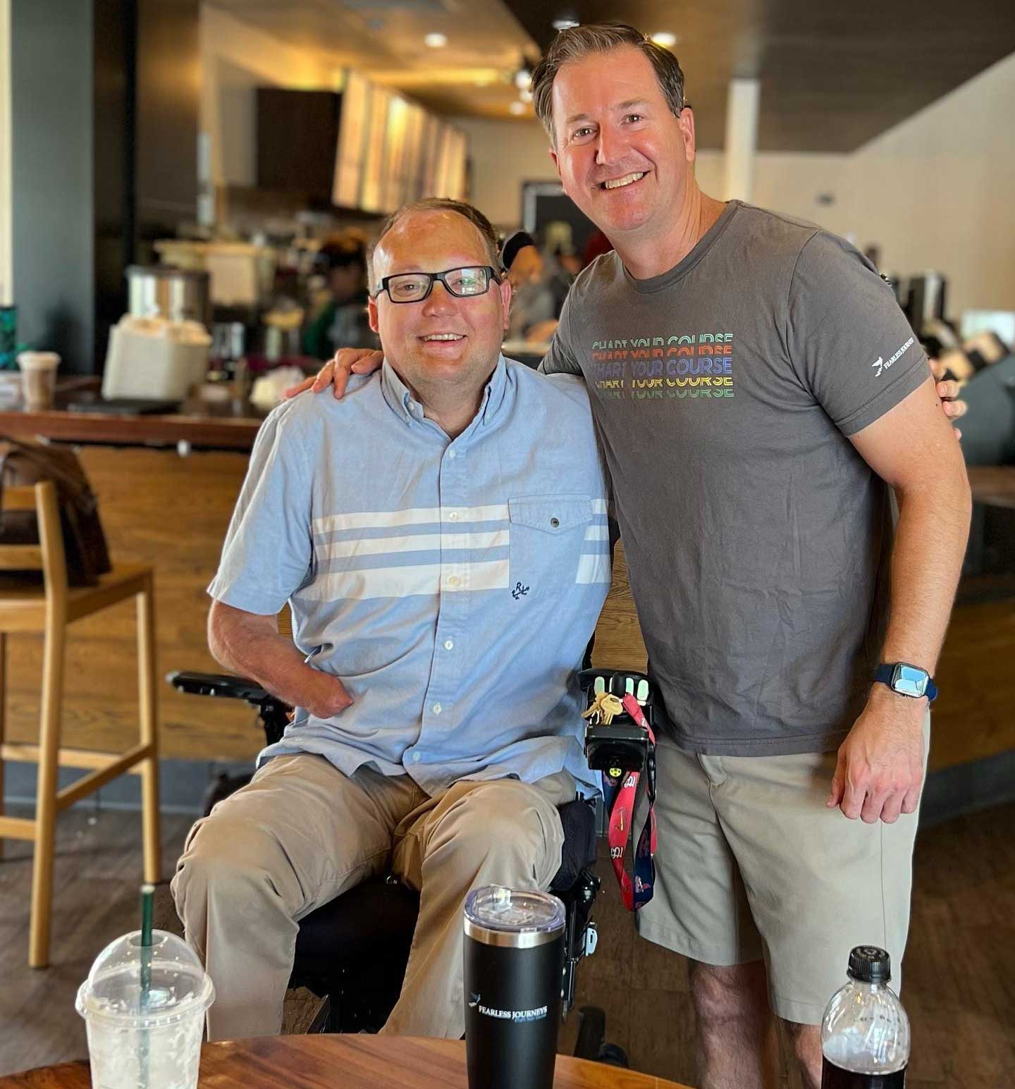 John seated in his wheelchair next to his friend at a Starbucks.