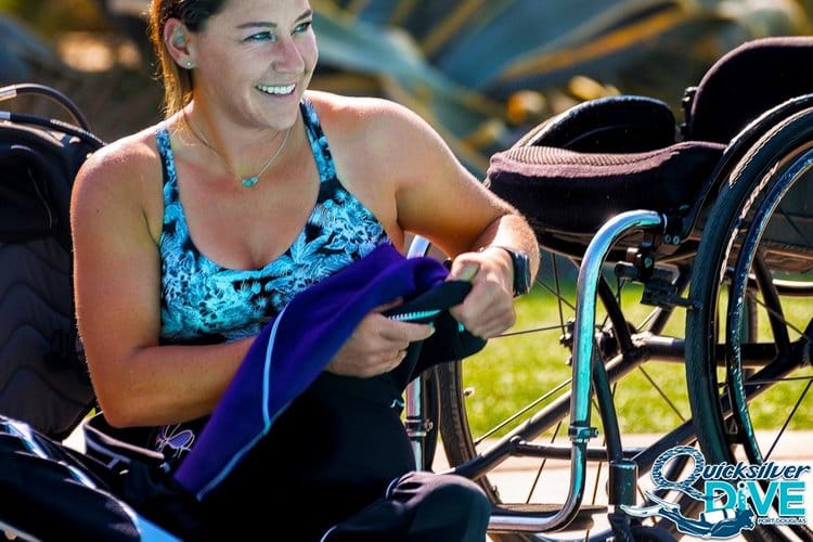 Disabled diver wearing a swimsuit seated on the ground next to her wheelchair.