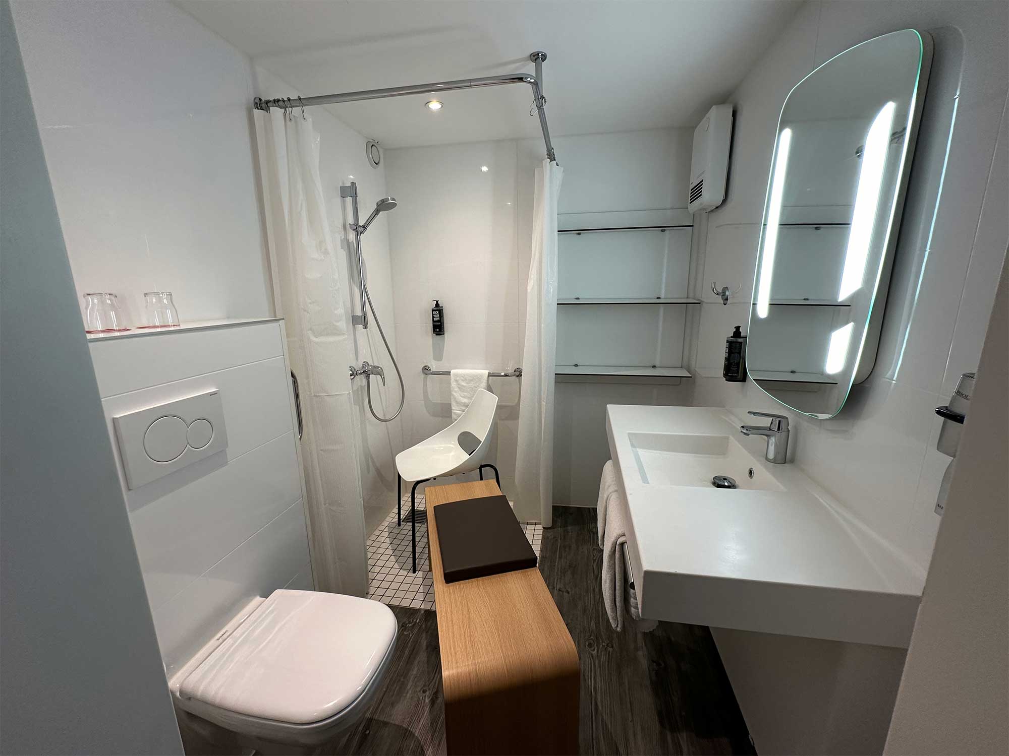 Hotel bathroom with a long wooden bench placed between narrow space and a plastic desk chair in the shower.
