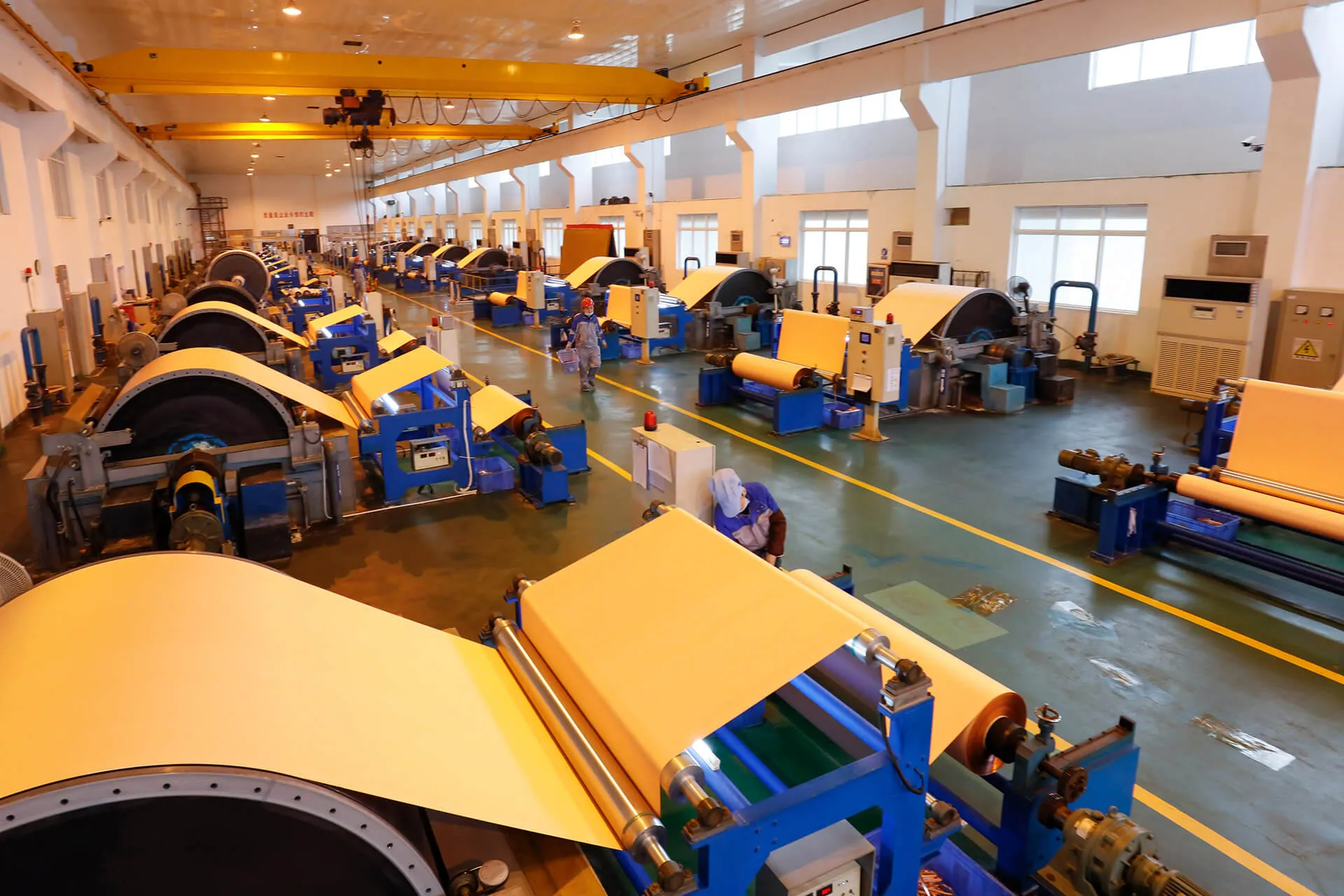Copper-colored foil sheets passing over rows of manufacturing drums and rollers in an industrial hall.