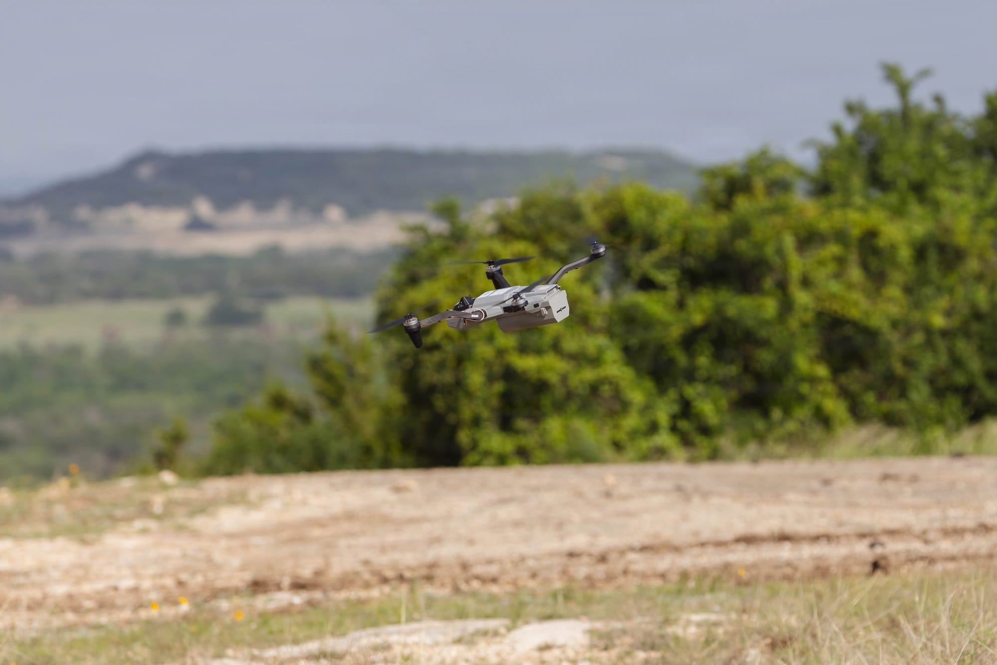 A Black Widow quadcopter banks in flight above a dirt clearing, with trees and rolling terrain behind it.