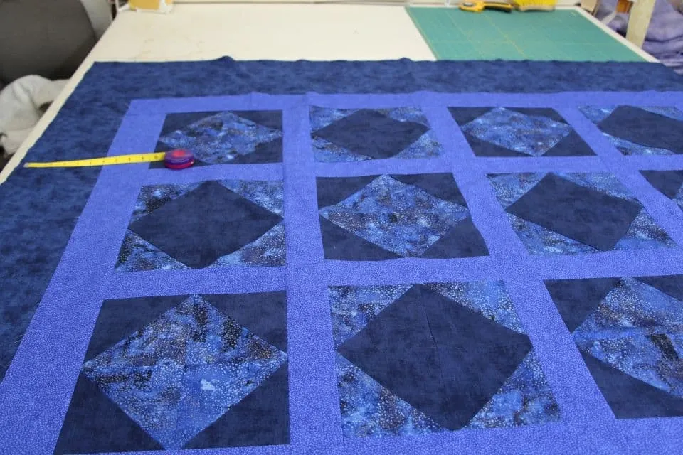 Dark blue quilt backing on the bottom layer where the yellow ruler lays depicting the 6 inches needed on all side of the quilt top which has blue diamond shapes and light blue marbled dots patterns