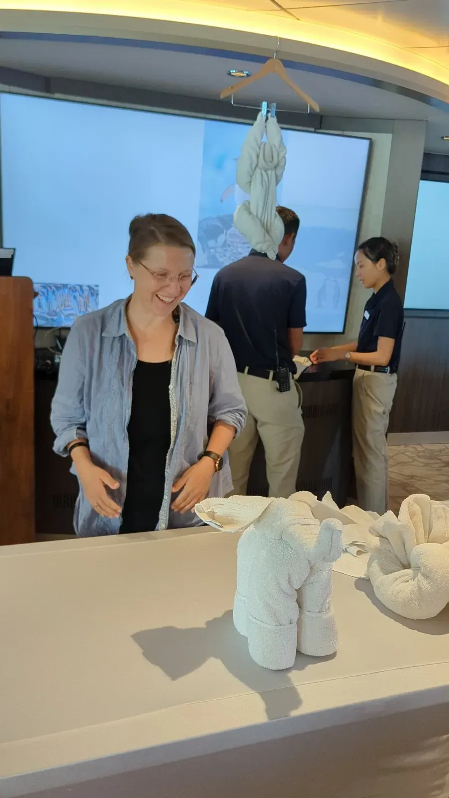 Katja lacht beim Handtuch-Workshop an Bord, vor ihr ein gefalteter Elefant aus Handtüchern, im Hintergrund ein Handtuch-Äffchen am Kleiderbügel
