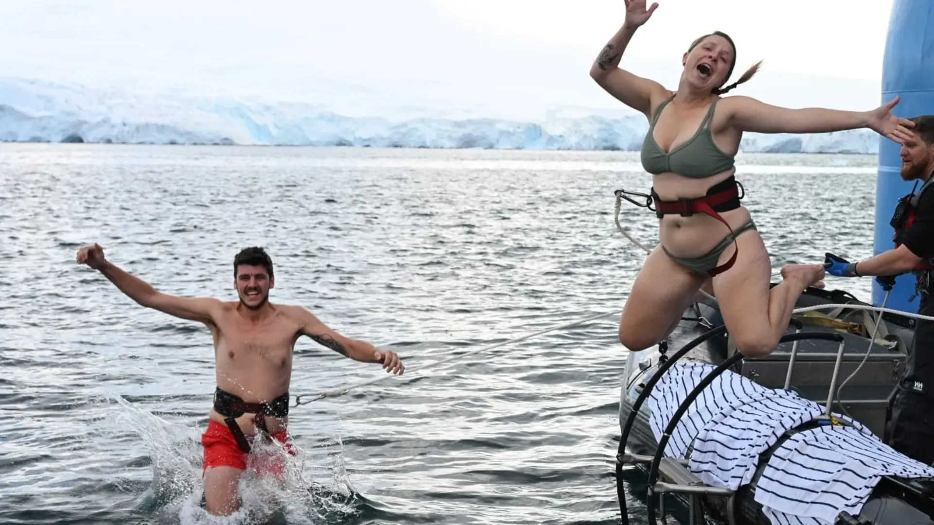 Katja und Nicolas springen angeseilt ins eiskalte Wasser der Antarktis, im Hintergrund eine Gletscherfront