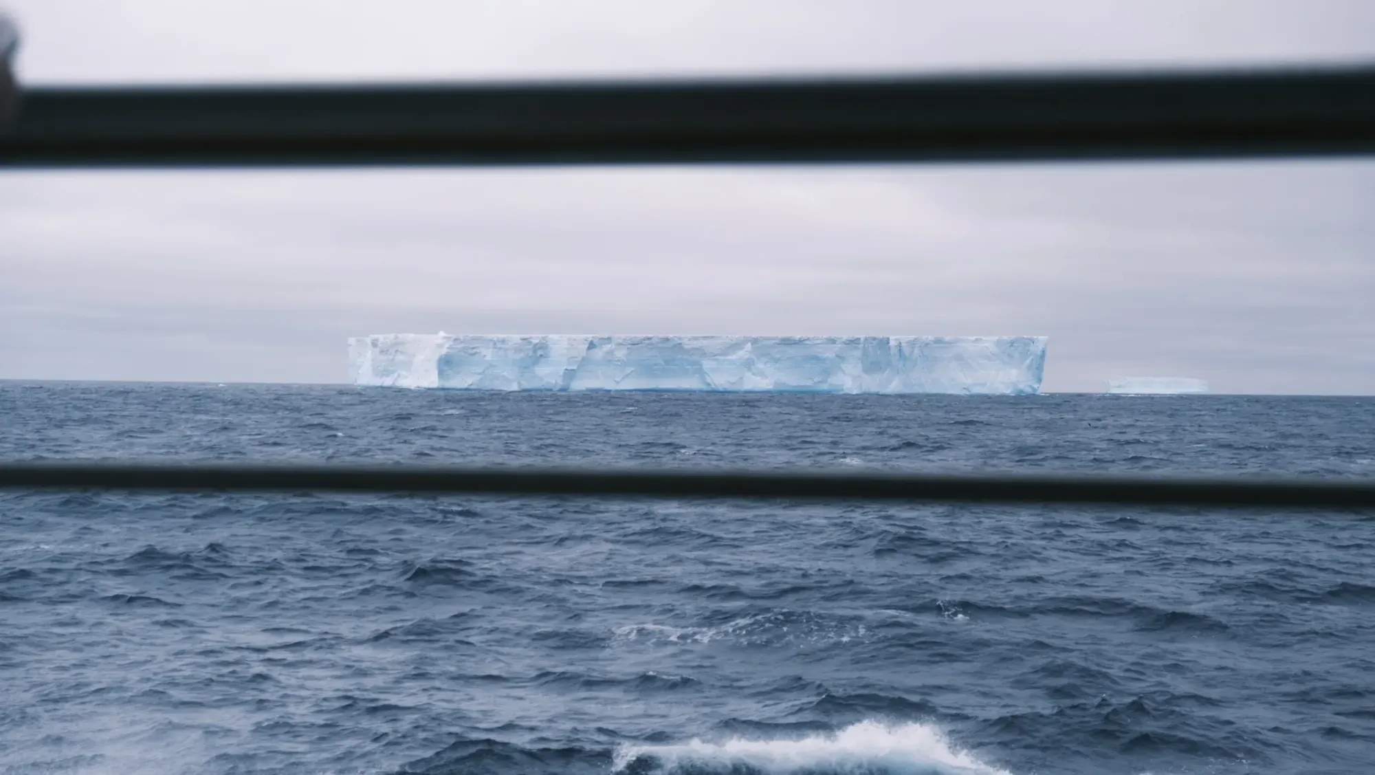 Grosser Tafeleisberg am Horizont, fotografiert durch die Reling des Expeditionsschiffs, im Vordergrund rauer Seegang