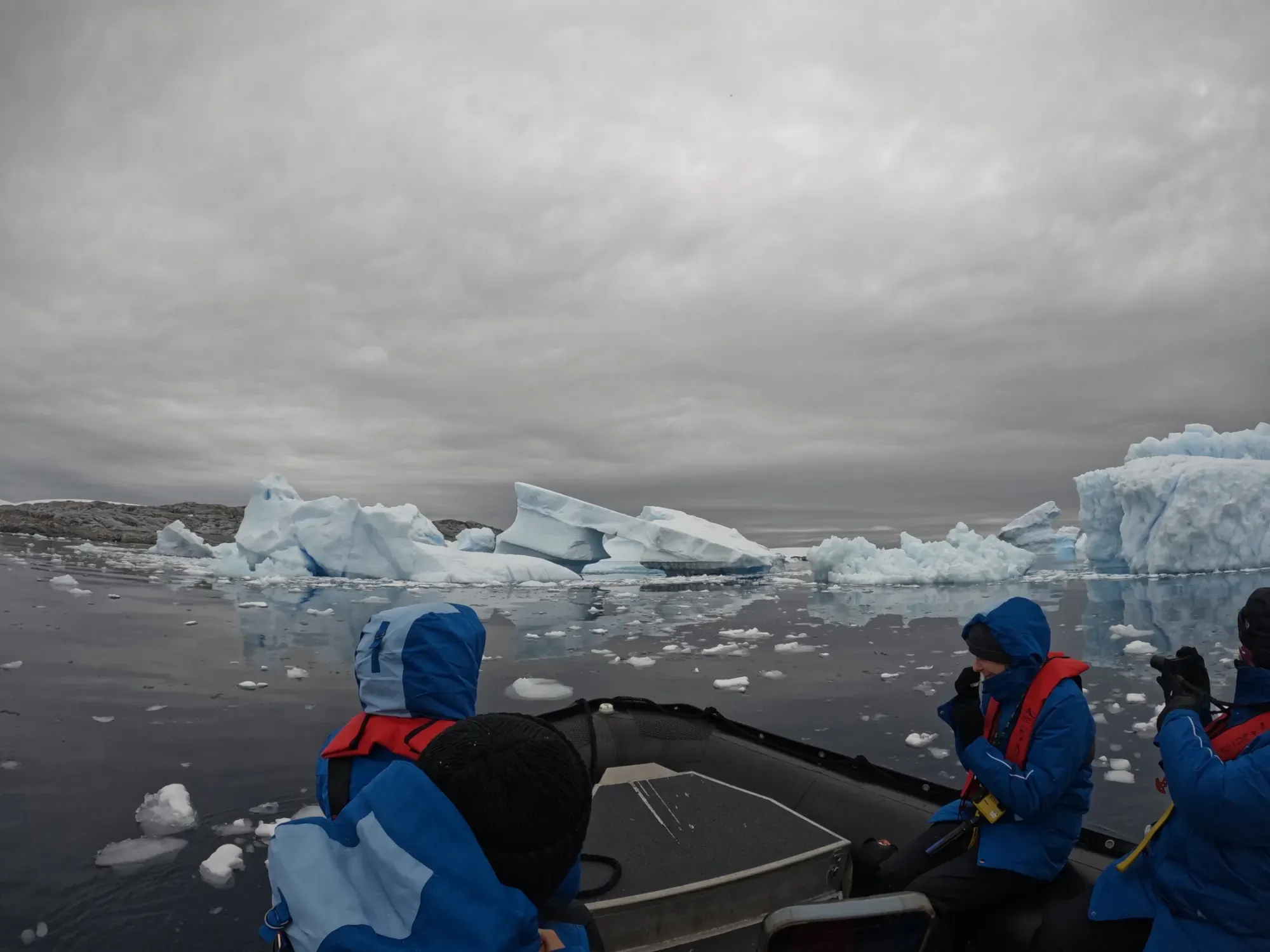 Gäste in blauen Expeditionsparkas und roten Schwimmwesten sitzen in einem Zodiac zwischen Eisbergen auf spiegelglattem Wasser