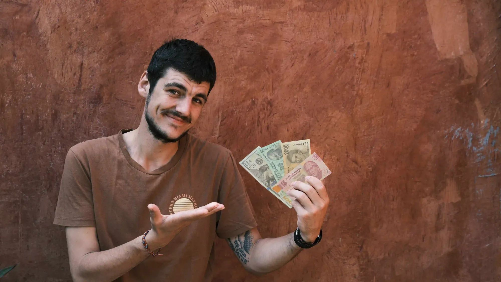 Ein junger Mann, welcher frontal in die Kamera schaut und mit seiner rechten Hand auf verschiedene Geldscheine in seiner linken Hand zeigt