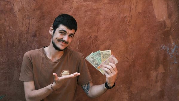 Ein junger Mann, welcher frontal in die Kamera schaut und mit seiner rechten Hand auf verschiedene Geldscheine in seiner linken Hand zeigt