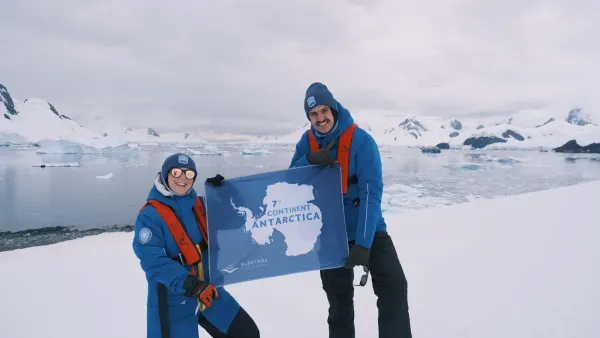 Katja und Nicolas in Expeditionsjacken halten eine Flagge mit der Aufschrift 7th Continent Antarctica