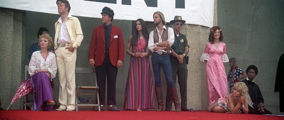 A group of characters in the film Nashville stand on a stage, watching a performance off camera.