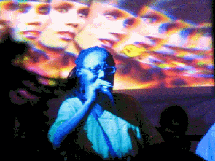 Cast in blue light, an older woman wearing eyeglasses and a neon green top performs in front of an animated GIF projection featuring Grace Jones.