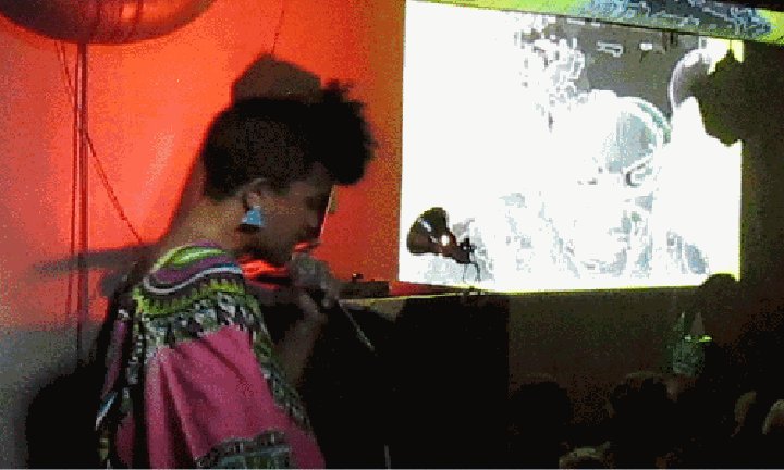 A female singer in a pink dashiki top sings to a crowd, standing in front a GIF projection, from "Sheroes #12: Nina Simone" (July 2020).