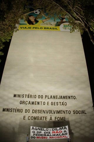 Protesters hold their banner in front of a government ministry building, which happens to have a soccer ad hanging at the top (courtesy Suyan Mattos) (click to enlarge)