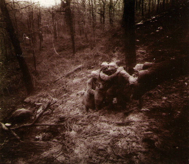 Two soldiers help a friend that had been shot. One dead body lay in the background.
