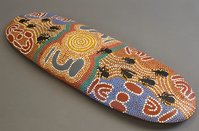 A beantree wood shield from Yuendumu, Northern Territory by Paddy Japaljarri Stewart, featuring a dot-style design in acrylic paint on the front. The artist, Paddy Japaljarri Stewart, is kirda (owner) of the story it shows.