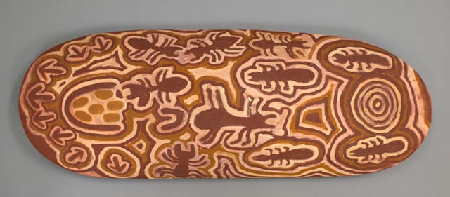A beantree wood shield from Yuendumu, Northern Territory by Paddy Japaljarri Stewart, featuring a dot-style design in acrylic paint on the front. The artist, Paddy Japaljarri Stewart, is kirda (owner) of the story it shows.