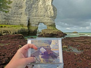 A researcher on the Texas State team holds a postcard of Monet's "La Porte d'Amont, Étretat" (c. 1868–69) in front of the present-day scene (via txstate.edu) (click to enlarge)
