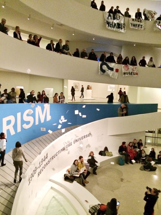 Flyers raining down onto the floor of the Guggenheim Museum in Manhattan, while protesters and chant and hold banners over the railings of the museum. (all photos by the author for Hyperallergic)