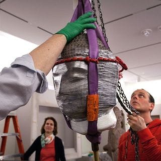 Museum Technician Michael Owen helps guide a sixth-century stone head of a Buddha, from China’s Northern Qi Dynasty, onto its new platform in the RISD  Museum’s Rockefeller Asian Galleries. Director of Planning Ann Woolsey, who has led the Museum’s extensive seven-year, $8.4 million Radeke Restoration Project, watches  with a smile. (Photograph by Erik Gould, RISD Museum Photographer. Courtesy of the RISD Museum,  Providence, RI.)