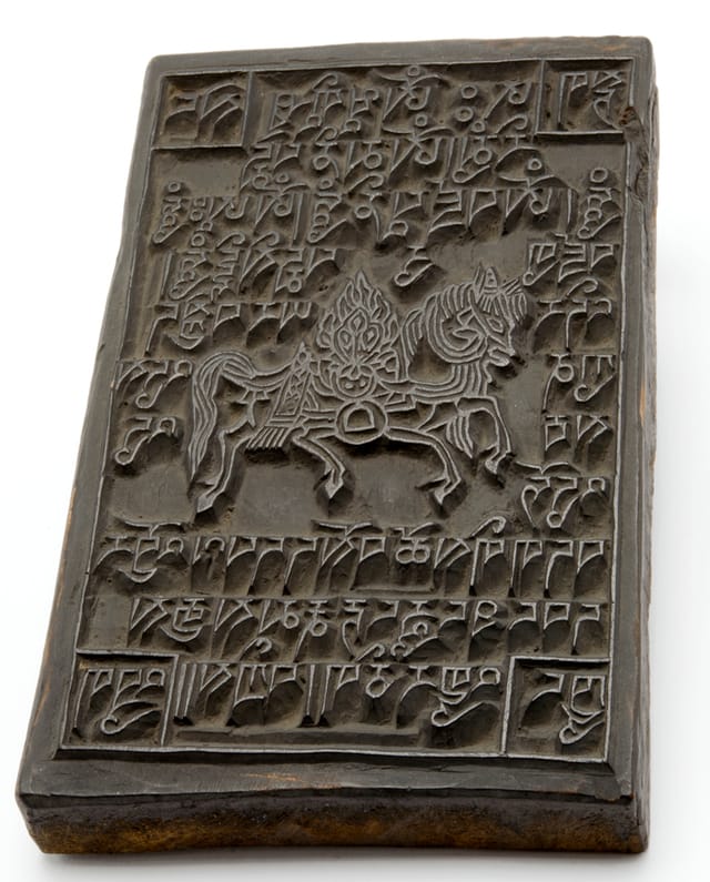 Printing block for prayer flag (lunta, rlung rta) Used to print the ubiquitous prayer flag with an image of a wind horse in the centre surrounded by mantras including om ma ni padme hum. In the four corners (clockwise from top left) are the names of powerful animals: tiger (stag), snow lion (seng), dragon (‘brug) and garuda (khyung). The text also includes the blessing words: ‘May [these] prayer flags increase merits, glory, richness and power [in your] life thanks to the lord! Fortune and Happiness!’ Wood. Height 19.6cm