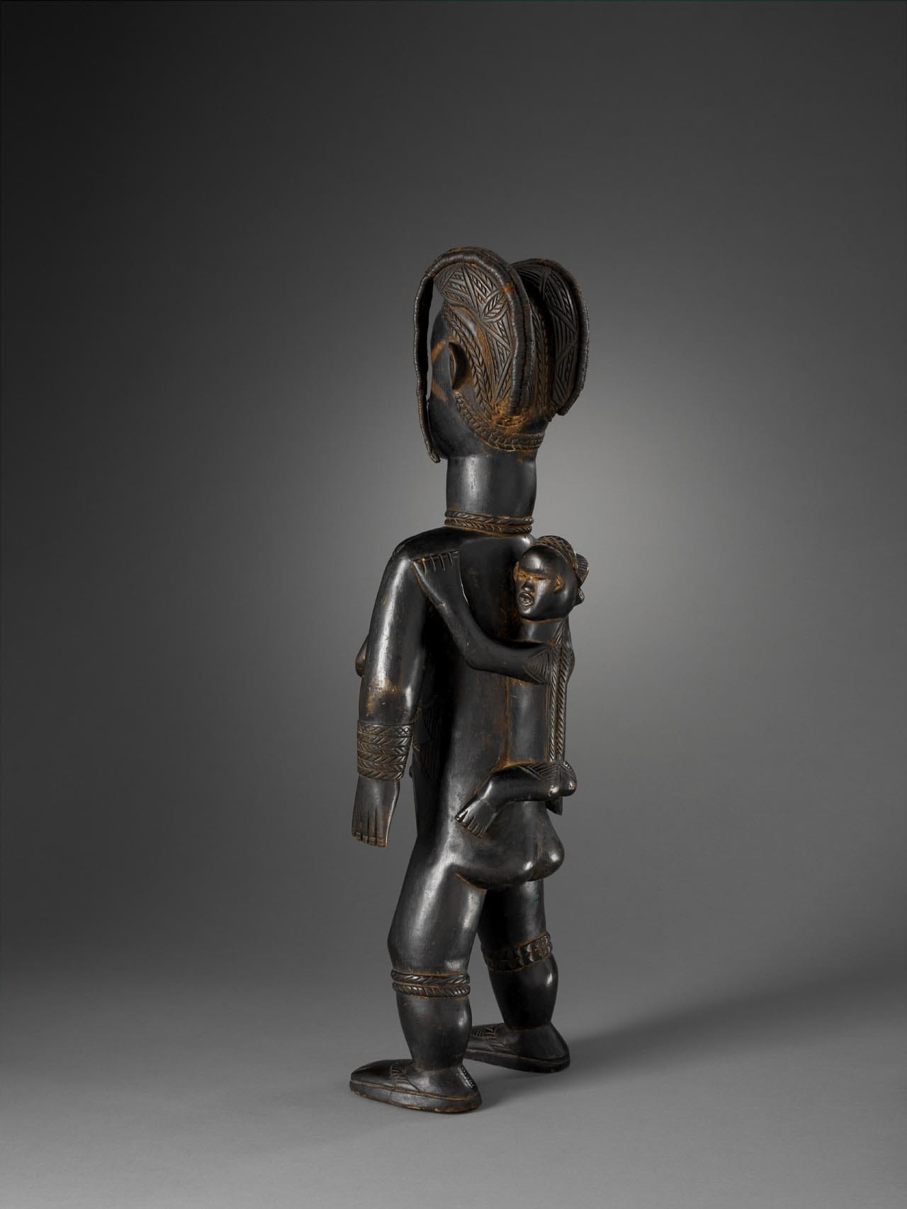 Statue féminine debout, bras le long du corps portant son enfant dans le dos. Le visage et le torse sont marqués de scarifications. Son visage est surmonté d'une coiffure en deux coques qui se prolongent par des mèches en fibres végétales tressées. Les yeux sont maquillés avec du kaolin. La bouche ouverte laisse voir des dents en métal. Elle porte des bracelets figurés au niveau des genoux et des mollets. Elle est chaussée d'une paire de sandales. Bois léger noirci, enduit rougeâtre sur les yeux. Haut. 63 cm, larg. 20,5 cm, prof. 16 cm