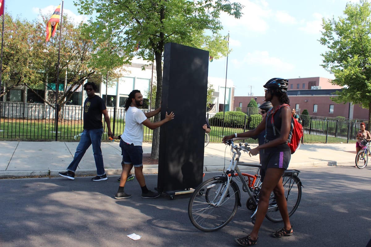 Danny Giles pushes his work slowly through the procession, a black monolith that offers a funny joke about the pretenses of public art and his presence in society as partly alien