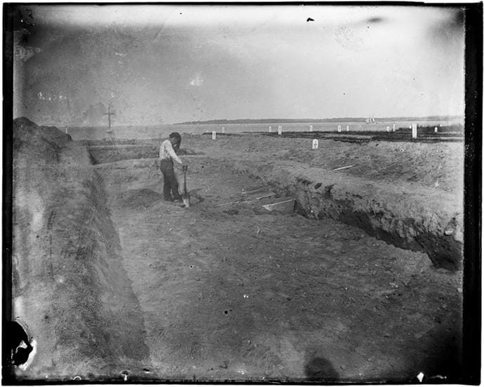 Jacob Riis, "The Potter's Field Consecrated Ground" (1890), gelatin dry plate negative. The image shows a man covering coffins with dirt in an open trench, a burial process mostly unchanged today. (courtesy Museum of the City of New York)