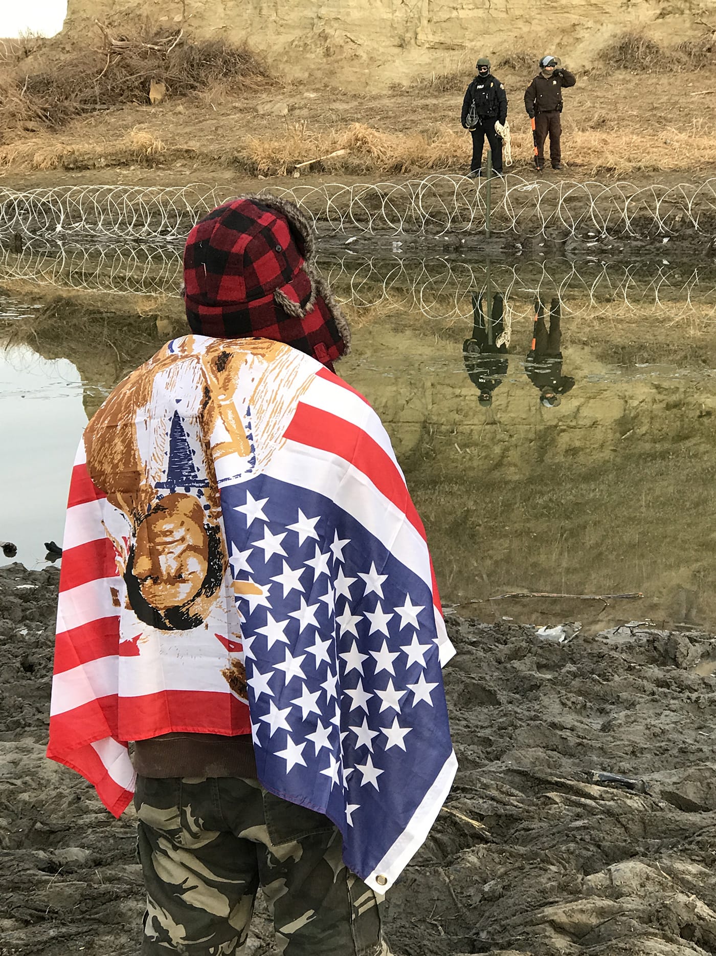A Native American protector facing police forces across the river who are installing barbed wire after taking over the area from protestors the night before.