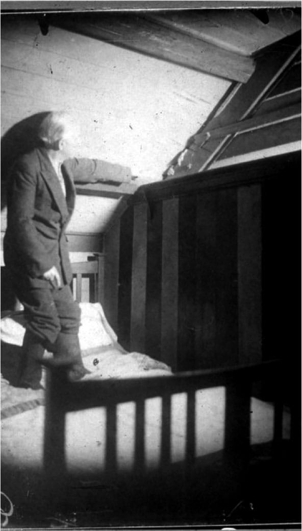 "'How Gef showed his fingers' — Irving is pointing towards a pair of white, hand-like objects that protrude from the gap between the wooden rafter and the sloping roof. Note that he appears to be wearing boots whilst clambering upon his bed!," from <em>Gef!: The Strange Tale of an Extra-special Talking Mongoose</em> (courtesy Strange Attractor)