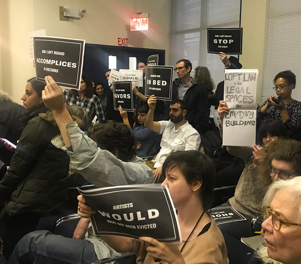 A public meeting of New York City's Loft Board on January 18, 2018 proceeds while protesters in the audience brandish signs.