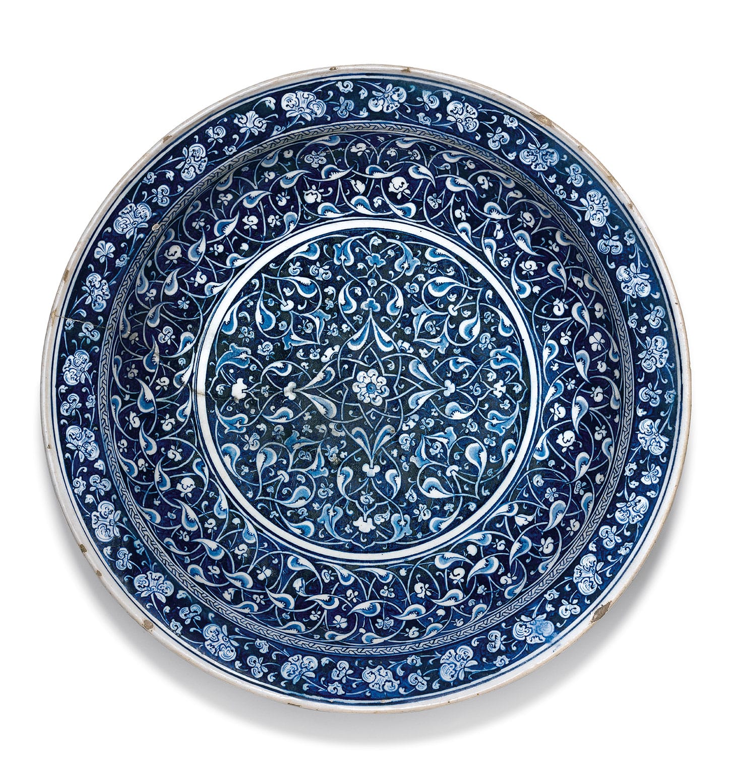 A highly important blue and white Iznik pottery charger, Turkey (c. 1480), of large, deep, rounded form, with an everted flat rim, the interior decorated in reserve within a central roundel against a blue-black ground with an intricate arabesque of interlacing split-palmette ‘rumi’ motifs in two shades of blue and white emanating from a small central rosette, the cavetto displaying a thick band of corresponding design surmounted by a thin band of interlocking key fret motifs, the flat rim ornamented with a band of scrolling floral ‘hatayi’ flowers in white, the reverse decorated with a wide band of ‘hatayi’ lotus scrolls in blue on a white ground, thin double blue lines and a single bracketed line embellishing the underside of the rim, 44.5 cm. diameter, 8.3 cm. depth (image courtesy Sotheby's)