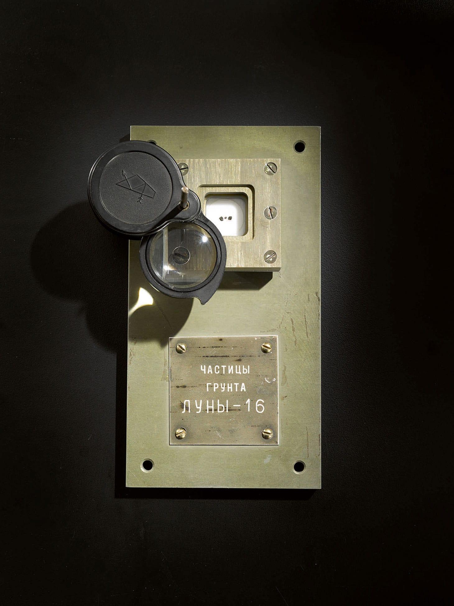 The only known documented samples of the moon available for private ownership, soil samples from Luna-16, collected September 1970, three samples of lunar rock from Luna-16, the first automated sample retrieval from the moon, encased under glass within a 2 by 2-inch metal block positioned below an adjustable lens, the whole secured to a 6½ x 3½ inch metal base labeled “ЧАСТИЦЫ ГРУНТА ЛУНЫ-16” [SOIL PARTICLES FROM LUNA-16], central fragment consisting of basalt with feldspar crystals visible, adjoining larger fragments consisting of finely structured vesicular agglutinates, showing glass coatings caused by age-old meteorite impact (image courtesy Sotheby's)