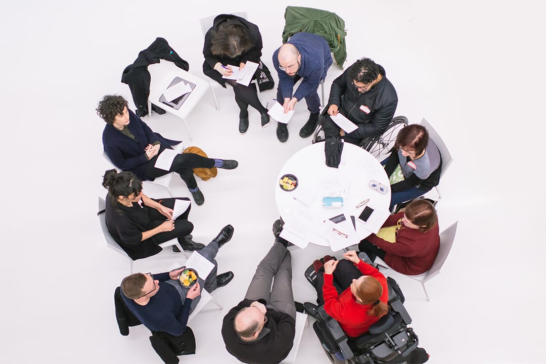 From above, disabled artists and activists form a circle around a table against a white floor. 