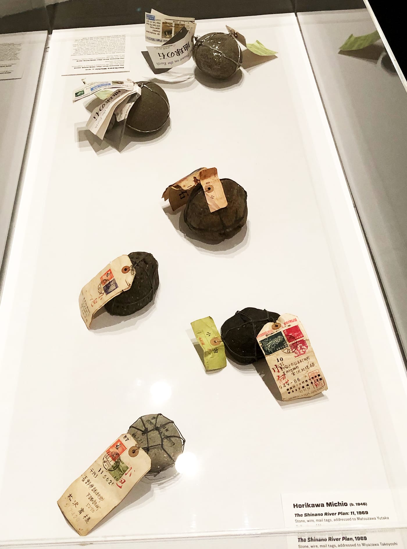 Michio Horikawa, works from various iterations of "The Shinano River Plan," a mail-art project that began in 1969; stones, wire, and paper tags; objects sent by postal mail and addressed to artists and other recipients (various collections, photo by the author for Hyperallergic)