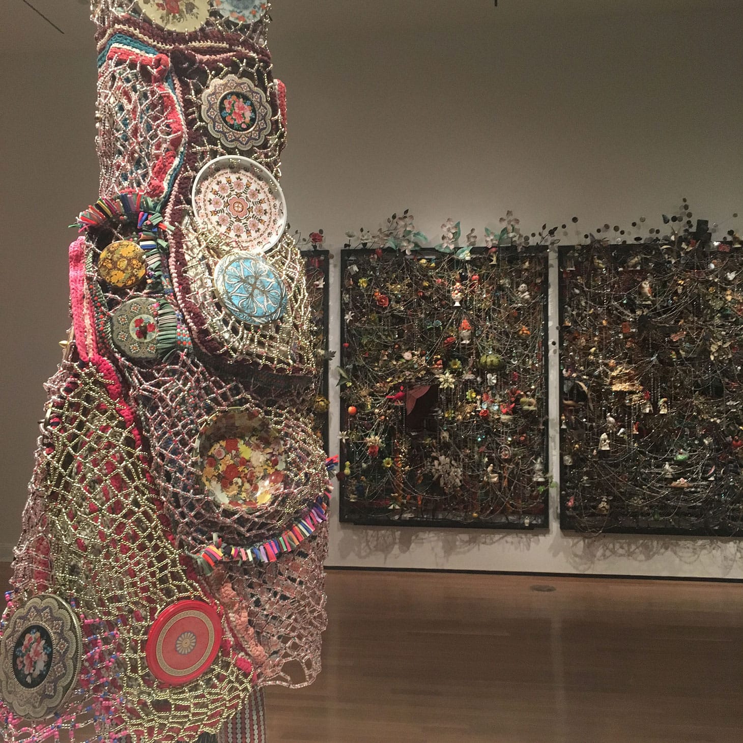 Nick Cave, detail of “Soundsuit” (2015), mixed media, including enameled can lids, macramé, shoelaces, plastic beads, metal, and mannequin in front of “Wall Relief” (2013), mixed media, including ceramic birds, metal flowers, afghans, strung crystals, and gramophone