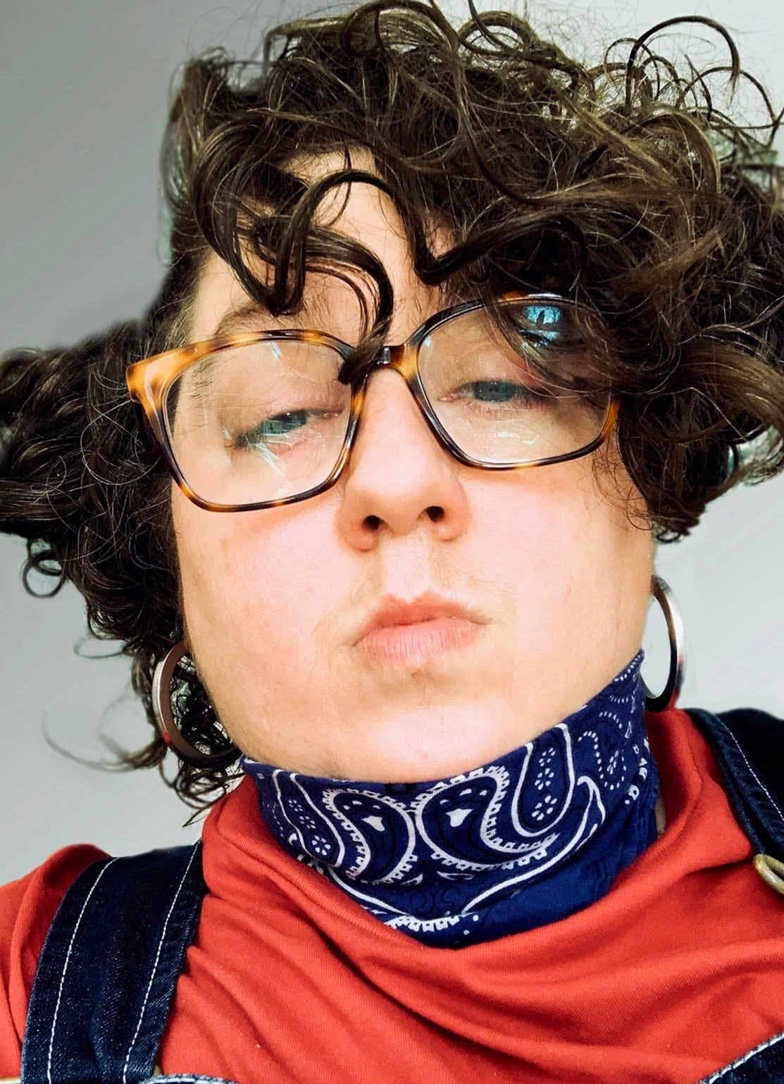Artist Constantina Zavitsanos (all images courtesy the artist); the artist wears a red top with a blue bandana around their neck, and glasses. They are looking down, towards the viewer, with their lips closed and curls framing their face.