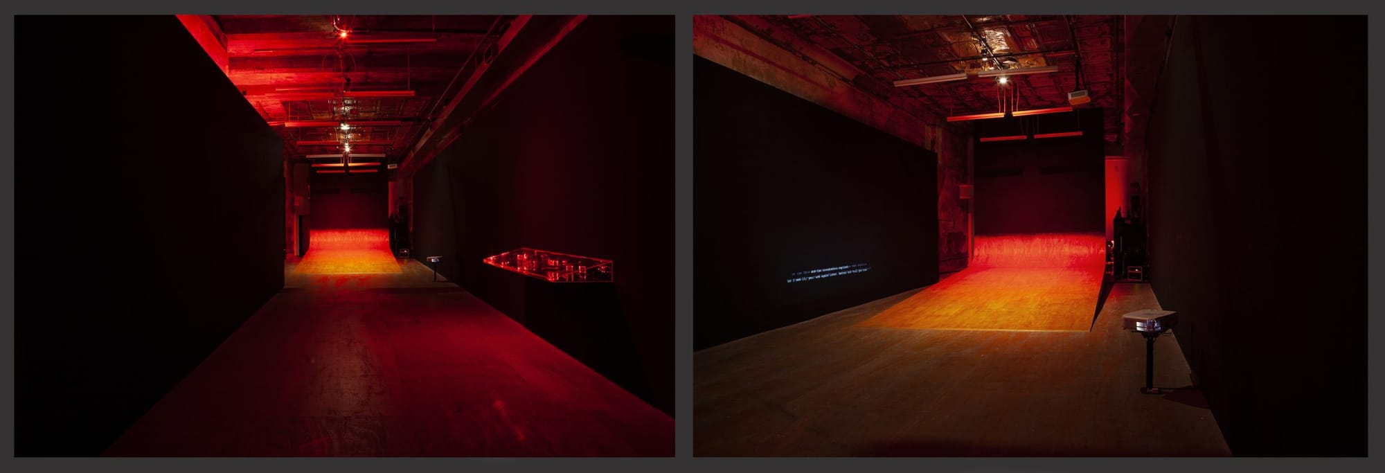 Views of a dark gallery space, filled with red light, host a large wooden ramp curling up the back wall. On the right is a mounted clear case glowing with red lasers; overlapping white and grey text is projected on the left wall.