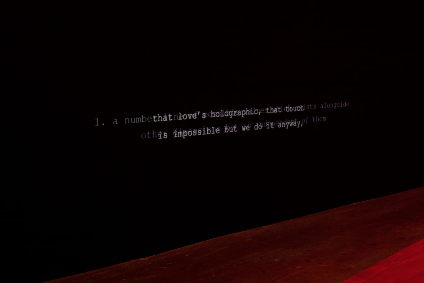 A black wall softly lit with red light shows two lines of white captions, reading: “that love’s holographic, that touch is impossible but we do it anyway” which overlap a second projection of illegible text.